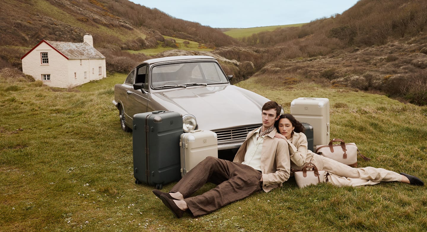 a man and woman sitting next to luggage on grass with a car